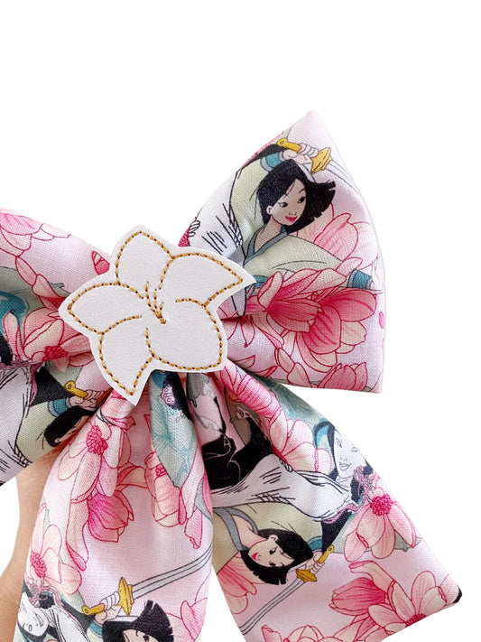 A close-up of a fabric hair bow featuring a pink and white floral print with cartoon characters, secured with a white flower-shaped hair clip.
